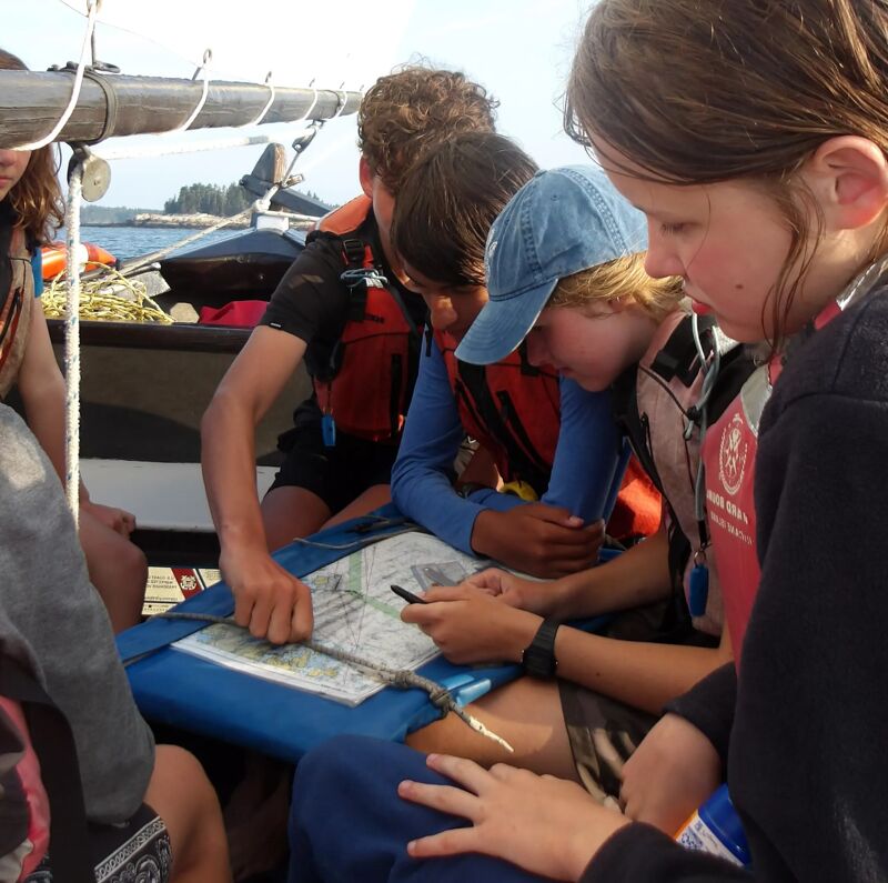 The photo shows a group of young people on a boat, possibly sailing. They are gathered around a map, seemingly navigating or planning their route. The individuals are wearing life jackets, suggesting a focus on safety. The background includes water and some land, indicating they are likely on a coastal or lake environment. The overall scene conveys a sense of teamwork and adventure.